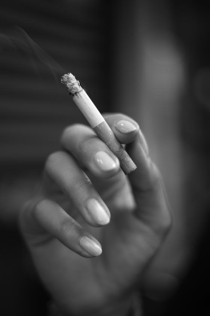 A close-up black and white photo of a hand holding a lit cigarette, with smoke visible.